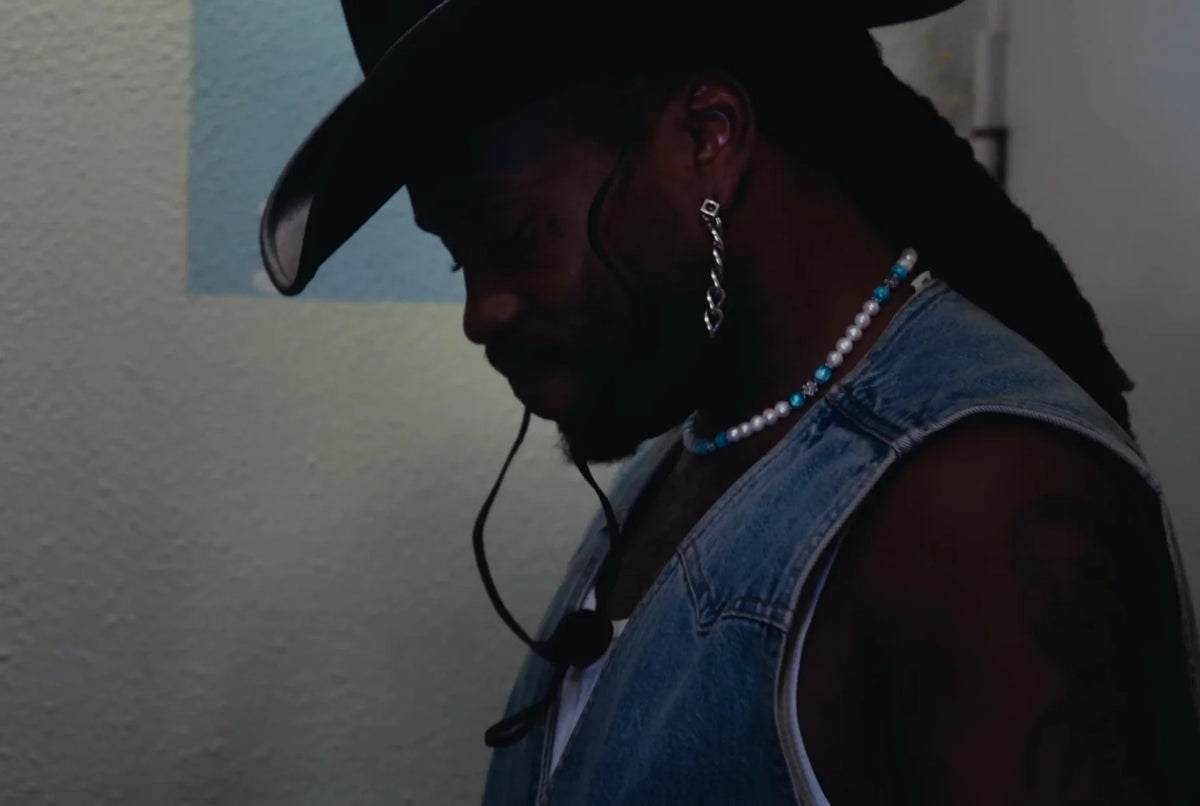 Jok’Air de profil, tête baissée, chapeau de cow-boy noir et veste en jean sans manches, Collier MIMOSA GLOW ICE au cou
